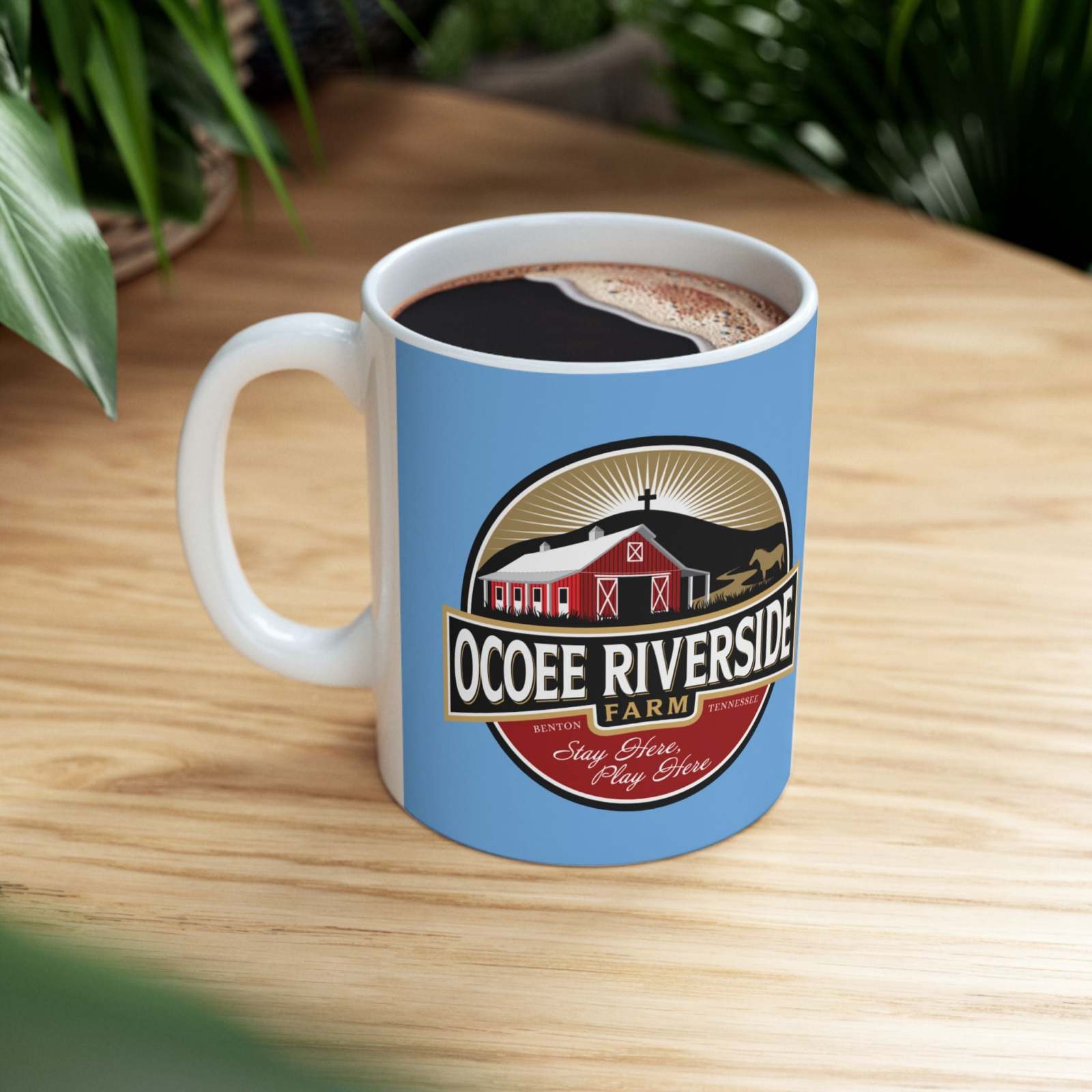 Glossy ceramic coffee mug with Ocoee Riverside Farm logo on light blue background filled with coffee on wooden table