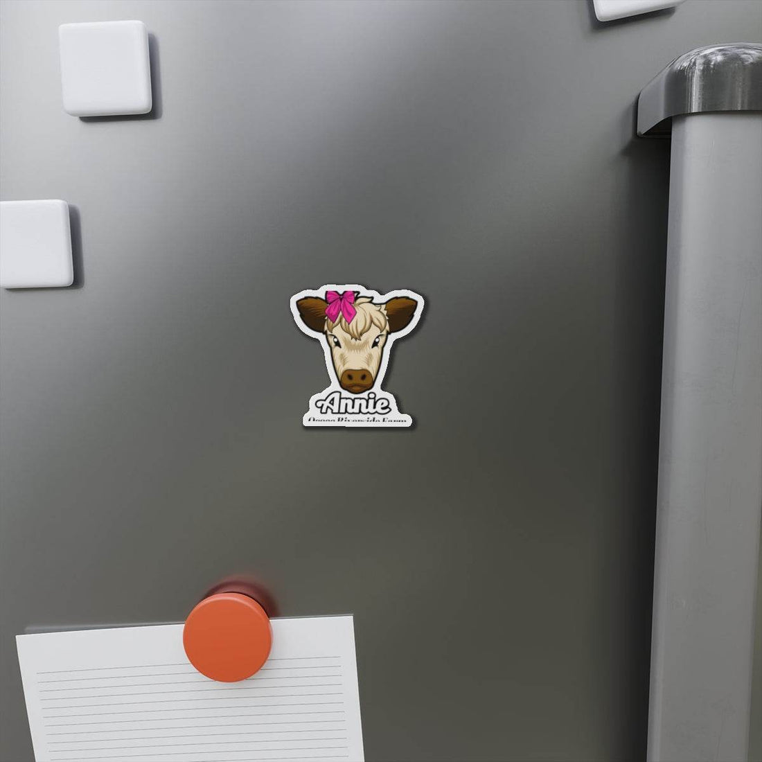 Cow die-cut magnet with a cute cow face and pink bow on a gray fridge holding a note with an orange magnet