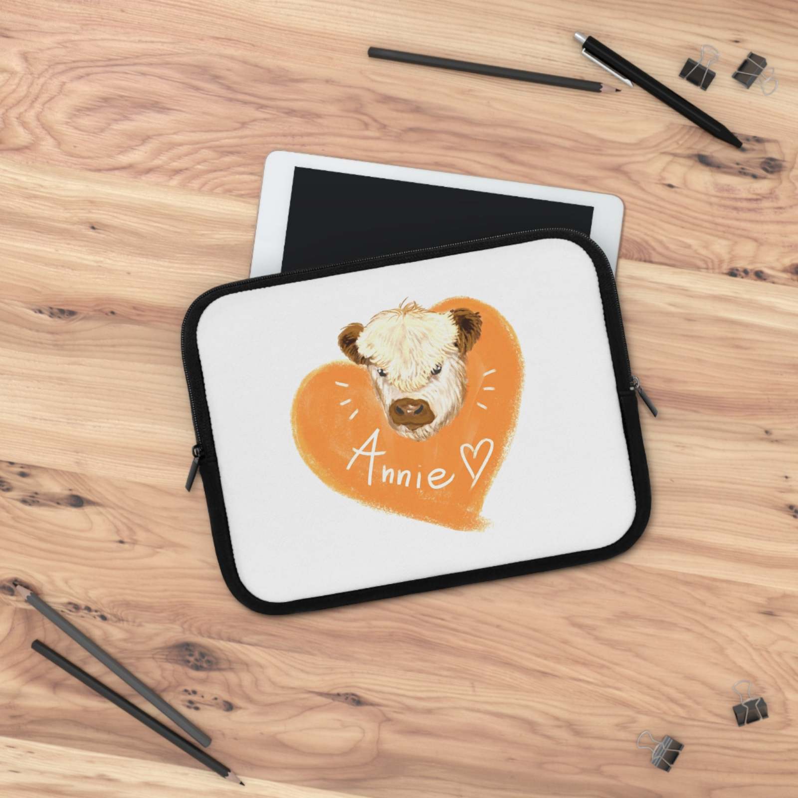 Custom Cow Laptop Sleeve with heart design and personalized name on a white neoprene case with black trim, displayed on a wooden surface with pencils and tablet nearby.