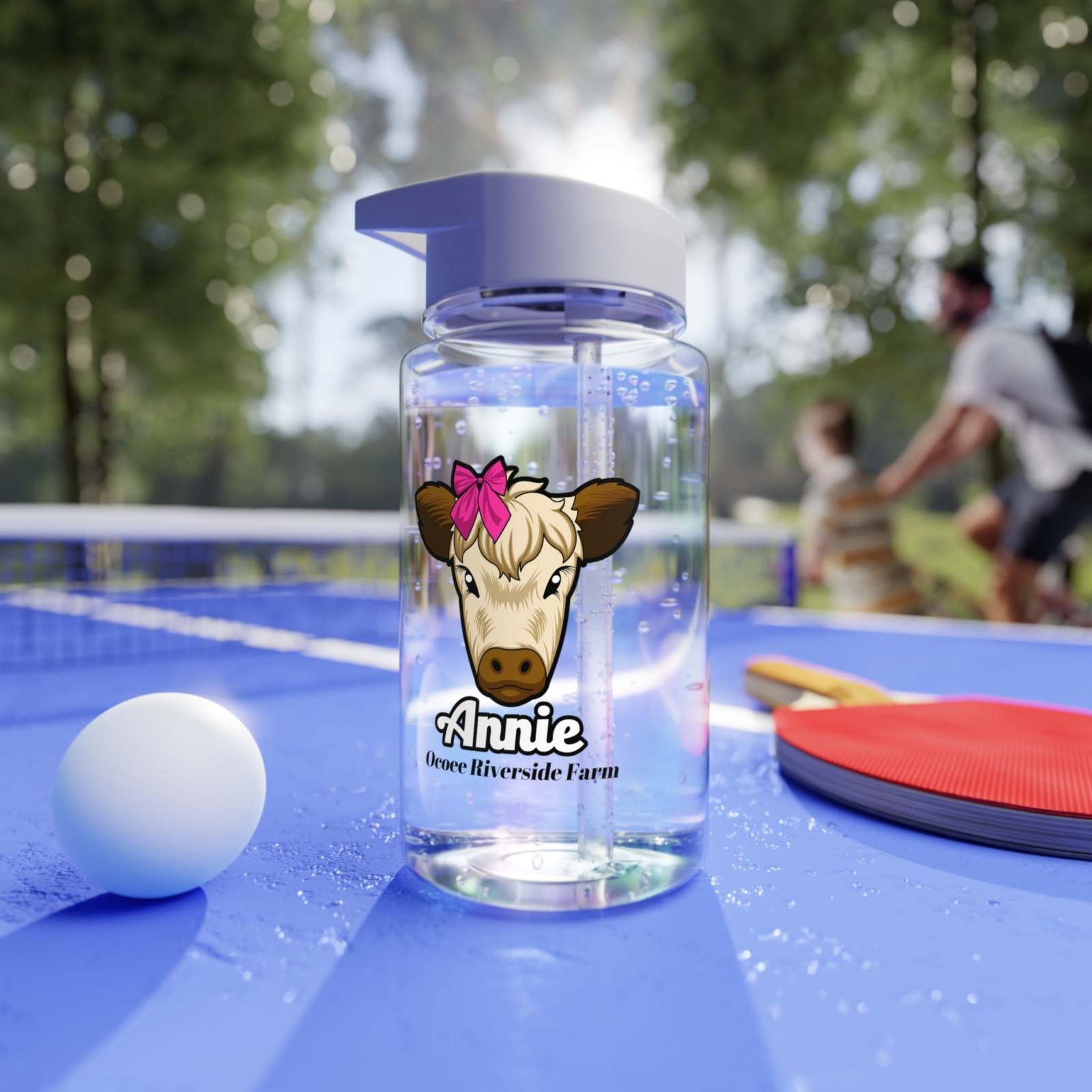 Clear Farm Animal Water Bottle with cow illustration and pink bow, featuring customizable name, on a blue sports table with ping pong paddle and ball outdoors
