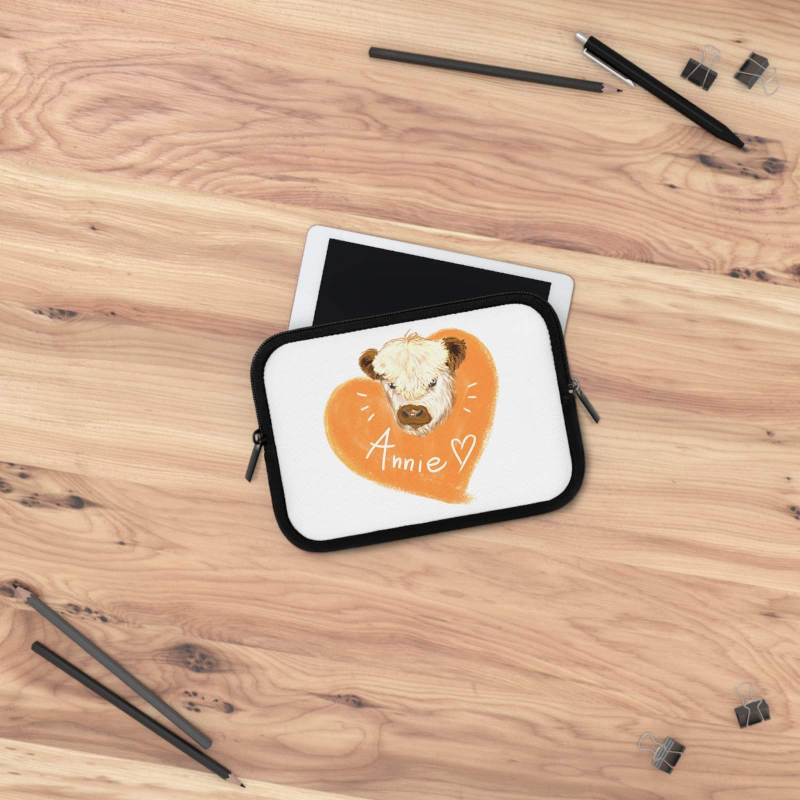 Custom Cow Laptop Sleeve with heart design and personalized name "Annie" on white neoprene, placed on wooden surface with tablet and black stationery items.