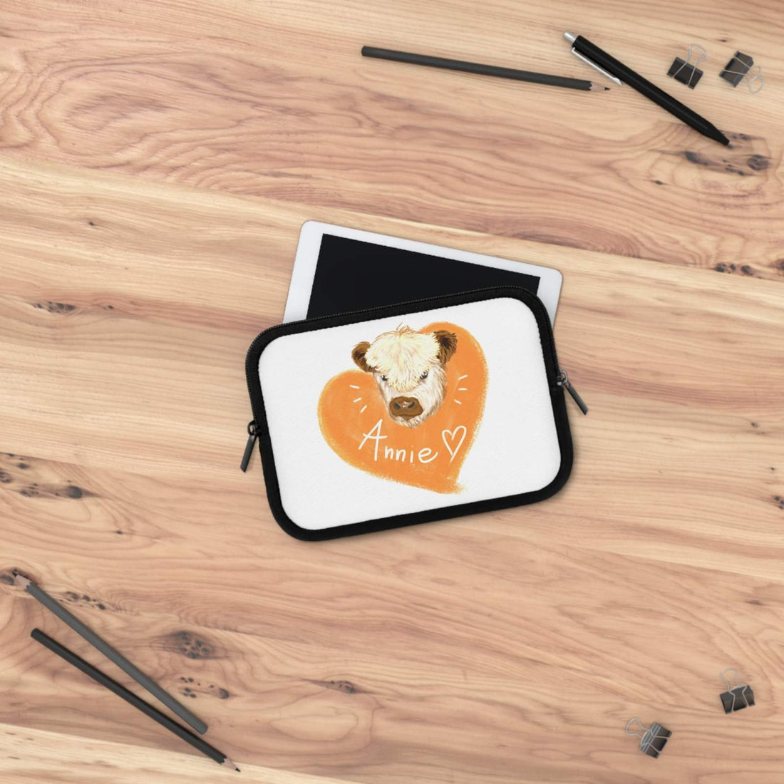 Custom Cow Laptop Sleeve with heart design and personalized name "Annie" on white neoprene, placed on wooden surface with tablet and black stationery items.