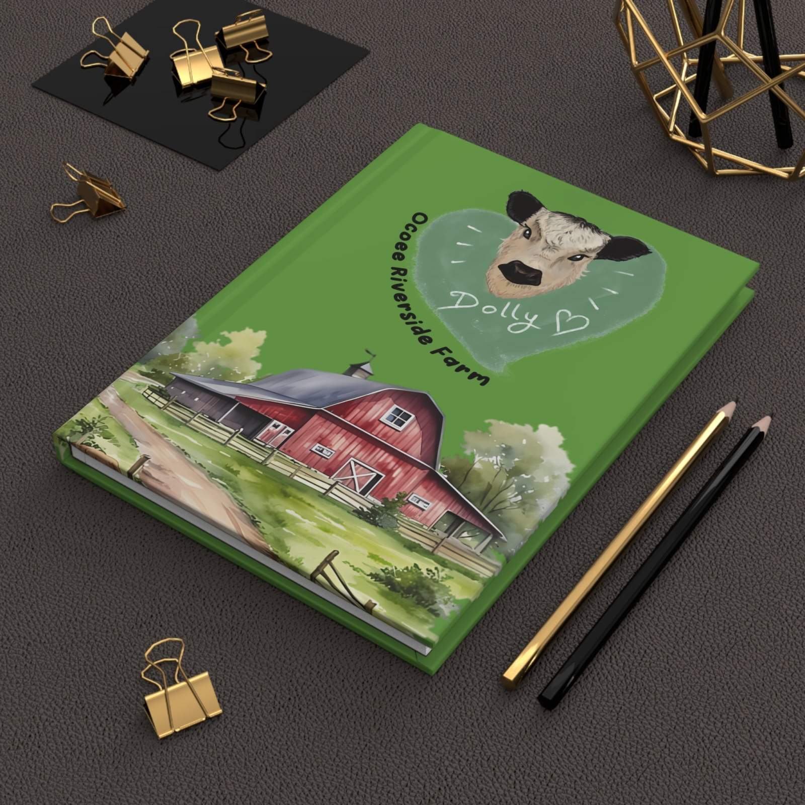 Country Farm-Themed Journal with green hardcover featuring a red barn and a sheep illustration, lying on a dark textured surface with pencils and gold binder clips nearby