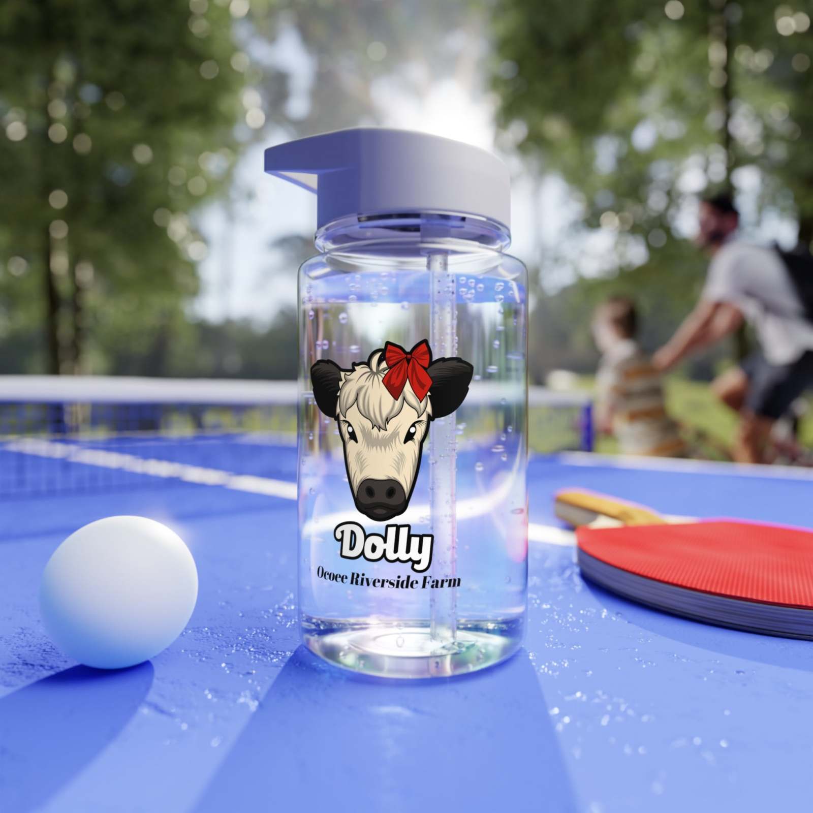 Clear cow print water bottle with straw lid featuring a cute cow face design, placed on a blue ping pong table outdoors.