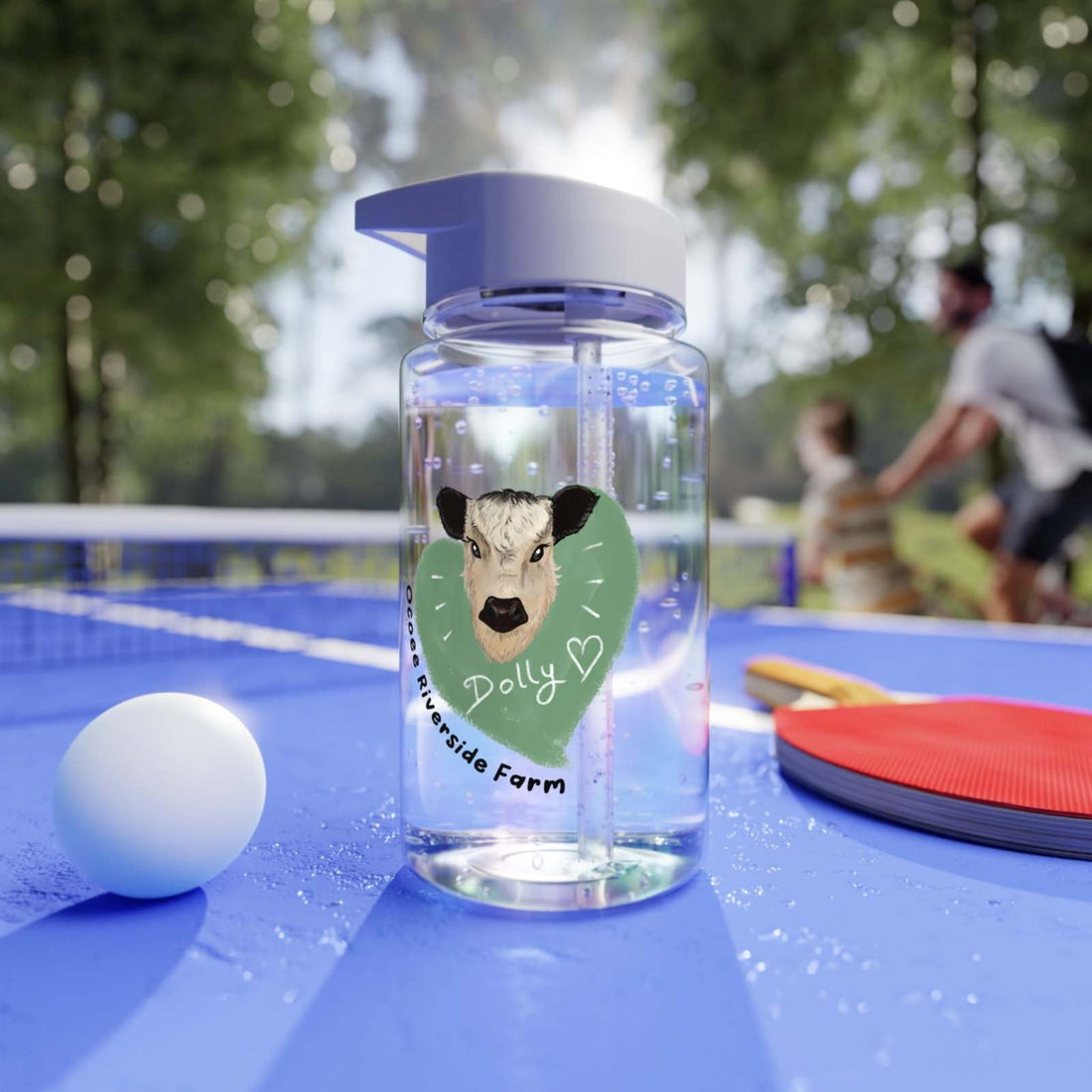 Clear farm-themed water bottle with a cow illustration and foldable straw, placed on a blue table tennis table with paddles and ball outdoors.